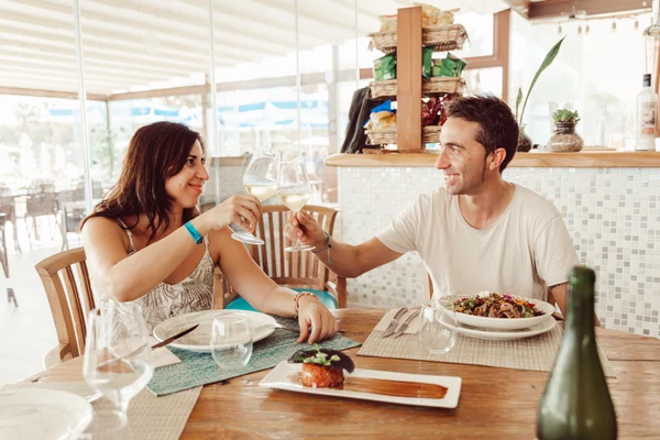 Two people clinking wine glasses, smiling across a table with food, in a bright, cozy restaurant setting at Taïga Delta De L'Ebre