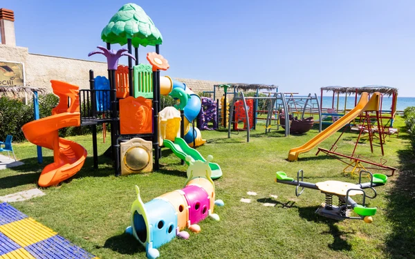 Colorful playground equipment sits on grass with slides and climbing structures under a clear blue sky near the sea at Triscina Mare