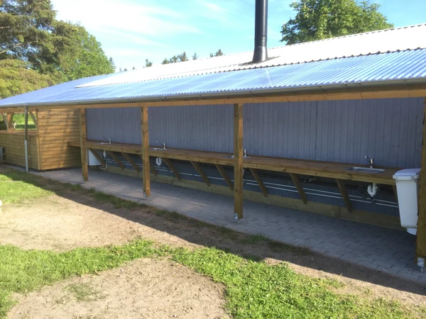 Wooden washing station with sinks and faucet under a roof, standing on a paved area with grass and trees nearby at Ballum