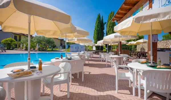 White patio tables with matching chairs under umbrellas, set near a pool, surrounded by greenery and buildings at Residence Eden