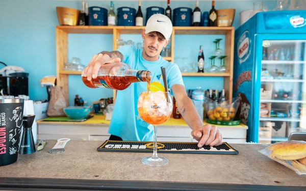 Bartender pouring orange drink into glass garnished with orange slice and straw, in a colorful bar setting at Triscina Mare