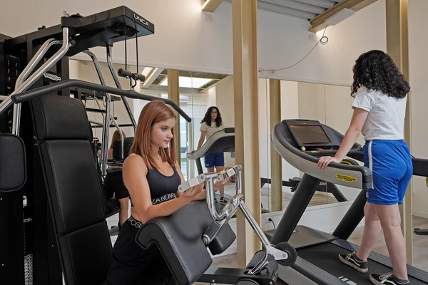 Women exercising with gym machines, one using a weight machine, the other walking on a treadmill, indoors at San Francesco