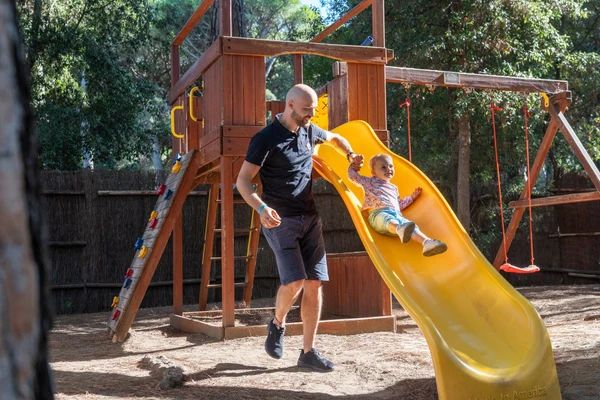 A child slides down a yellow slide while an adult assists in a wooded playground at Etruria Camping Village