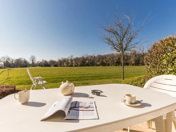 A white table holds tea items and magazine, set in a grassy park with chairs and leafless trees at Le Green Beach