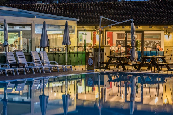 Poolside chairs and umbrellas reflecting in a calm pool surface; behind them, a café with people dining indoors at hu Venezia Camping in Town