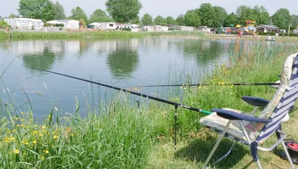 Fishing rods are set up by a lake, with a striped chair nearby, at a campsite surrounded by RVs and trees at Betuwe
