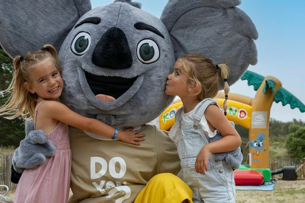 Two smiling children embrace a large koala mascot, set in a playful outdoor area with inflatable structures at Maguide