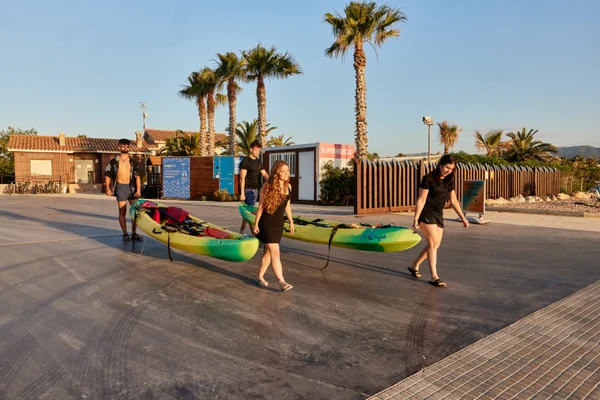 People pulling kayaks on a paved path, surrounded by palm trees and a building at Taïga Delta De L'Ebre
