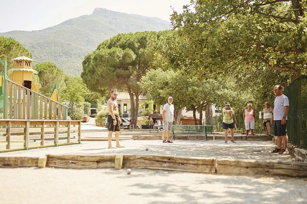 Group of people playing pétanque on a gravel court, surrounded by trees and a playground at Les Albères