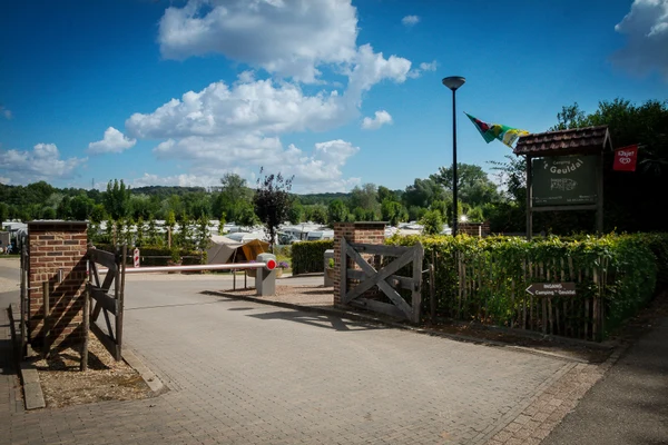 Entrance gate, partly open, leads to campsite with tents and caravans amidst trees and greenery, under a bright blue sky at 't Geuldal