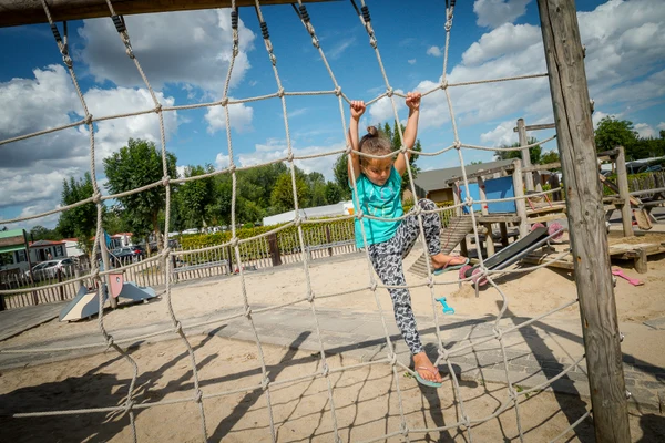 Child climbing rope net structure in sandy playground surrounded by wooden equipment and greenery at 't Geuldal