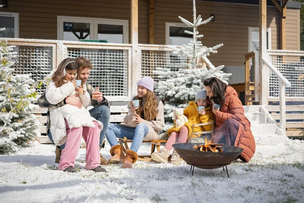 Family enjoying hot drinks around a fire pit, sitting on snow-covered ground near a wooden cabin at Marvilla Parks Kaatsheuvel