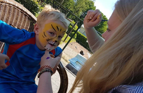 Child sitting while getting face painted as a tiger, outdoors near a fence and greenery at Hoeve de Schaaf