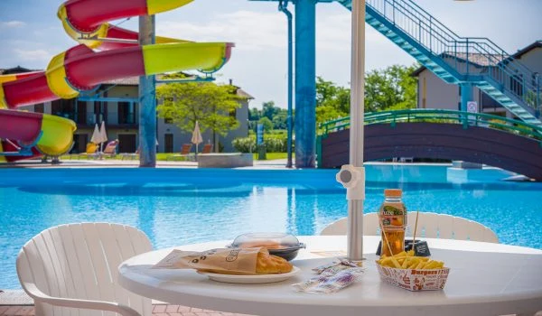 Plastic table with food and drink under umbrella, pool with slides in the background at Residence Eden