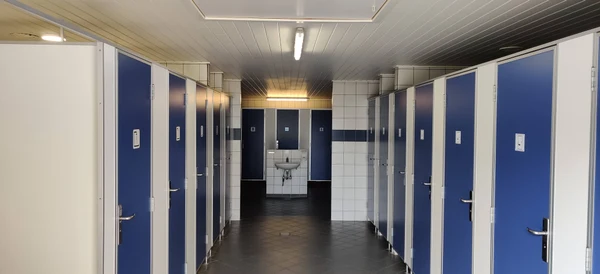 Multiple blue and white bathroom stalls facing a wall-mounted sink with mirrors, under fluorescent lighting at Ballum
