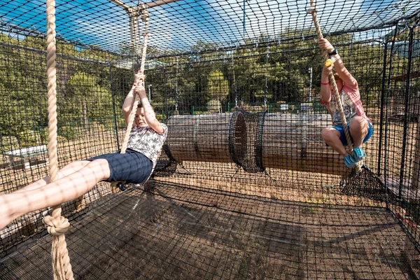 Two children swinging on ropes inside a netted play area, surrounded by a forest at Les Albères