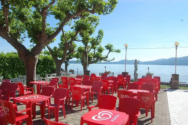 Red chairs and tables are arranged on a brick patio under shaded trees, overlooking a serene lakeside view at Lido di Monvalle