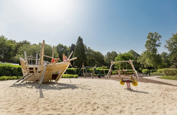 Children playing on a wooden ship structure in a sandy playground, surrounded by trees and additional play equipment at Hoeve de Schaaf