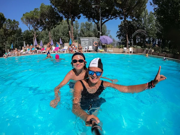 Two people smiling while swimming in a blue pool, surrounded by sunbathers and umbrellas at Punta Navaccia