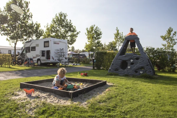 A child plays in a sandbox while another stands on a climbing structure; a camper is parked nearby at Camping de Molenhoek