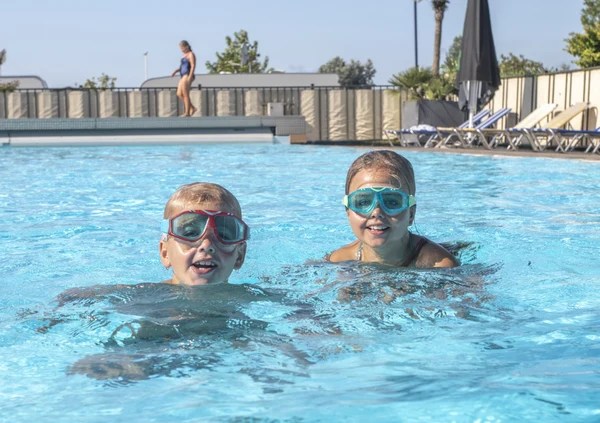 Children wearing goggles smiling while swimming in a clear blue pool, with lounge chairs and greenery nearby at Camping de Molenhoek