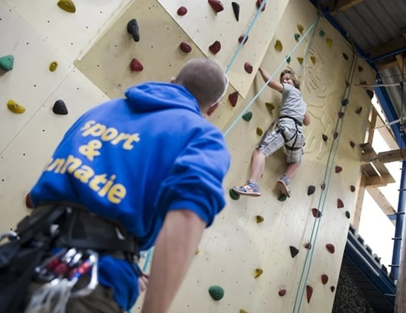 A child climbs a colorful indoor rock wall while an instructor in blue supervises at Hunzedal