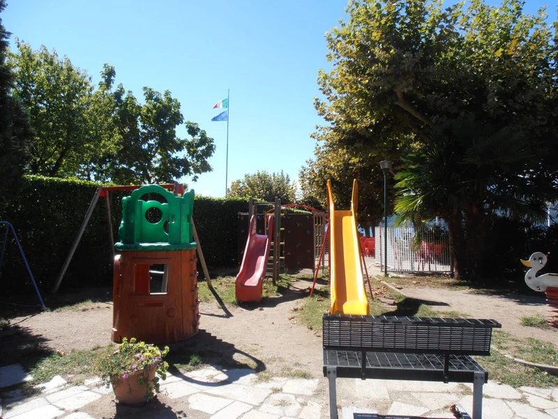 Children's playground features colorful slides and climbing structures amidst trees and a sunny, fenced park at Lido di Monvalle