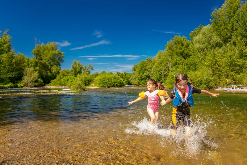 Two children splashing in a shallow river surrounded by lush greenery and clear skies at du Domaine d'Anglas