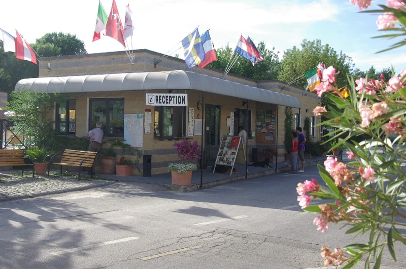 Reception building with people talking, benches, and potted plants, surrounded by flags on poles at Village Torre Pendente