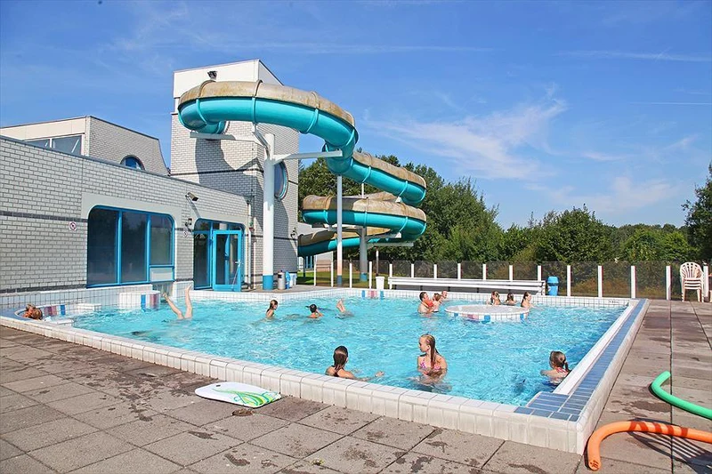 Children swimming near blue slides in a large rectangular outdoor pool on a sunny day at Hunzedal