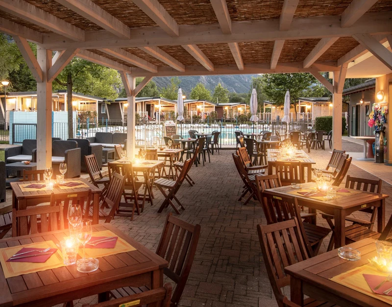 Wooden tables with candles arranged under a large, covered patio near a brightly lit pool area at Glamping II Borgo delle Arti