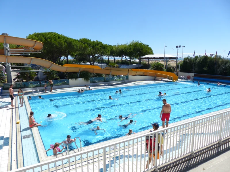 Swimmers enjoying a large, sunlit pool with water slides, surrounded by trees and seating at Internazionale