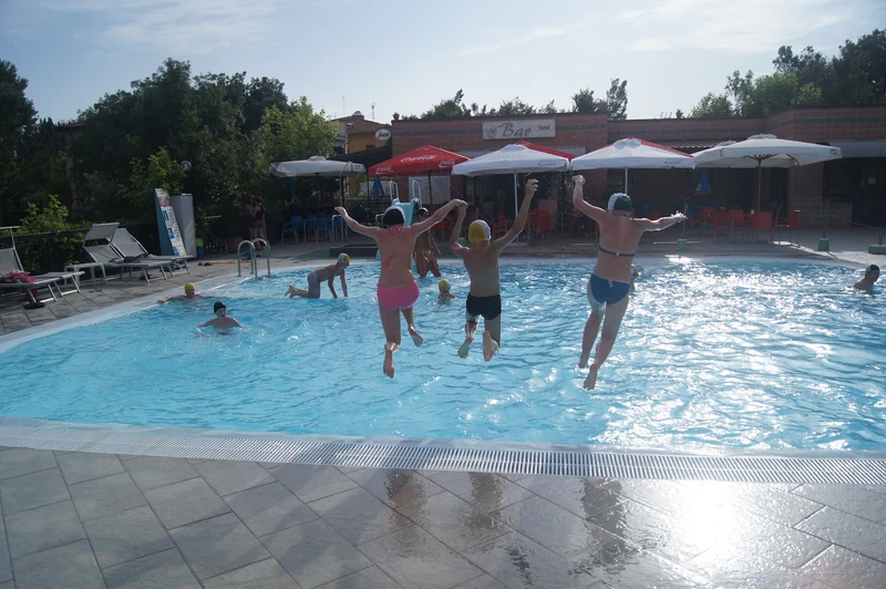 Four people leap into a swimming pool beneath umbrellas and trees at Village Torre Pendente