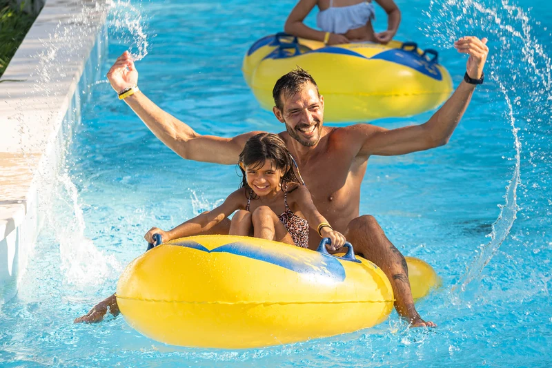 A man and child joyfully ride a yellow inflatable raft in a vibrant, sunny pool setting at Marina Julia Family Camping Village