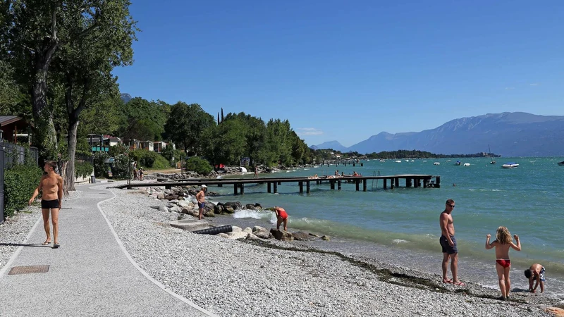 People walking, swimming, and relaxing along a rocky lakeshore with docks, surrounded by trees and mountains in the background at Villenpark Sanghen