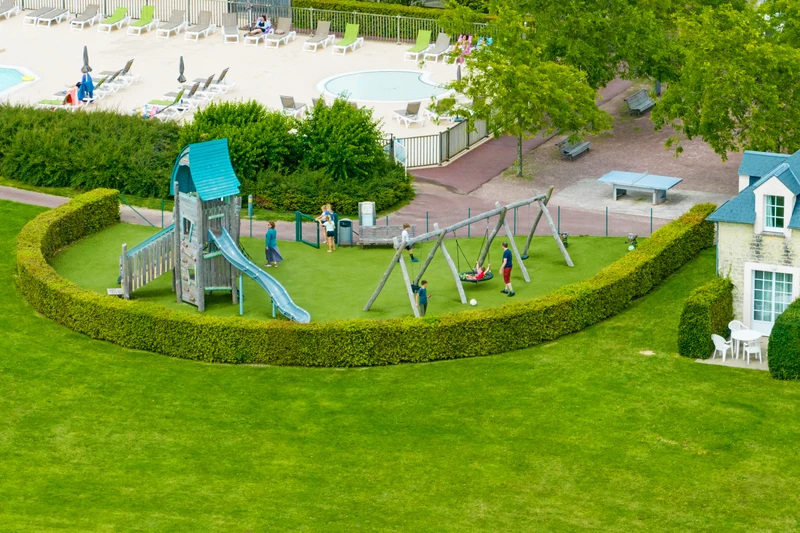 Children playing on a playground with slides and swings, near a pool and lounge chairs, surrounded by hedges at Le Green Beach