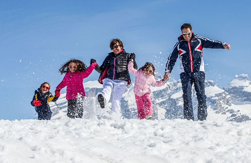 Family jumping joyfully, holding hands, surrounded by snow on a sunny day with mountains in the background at Cristal Palace Resort