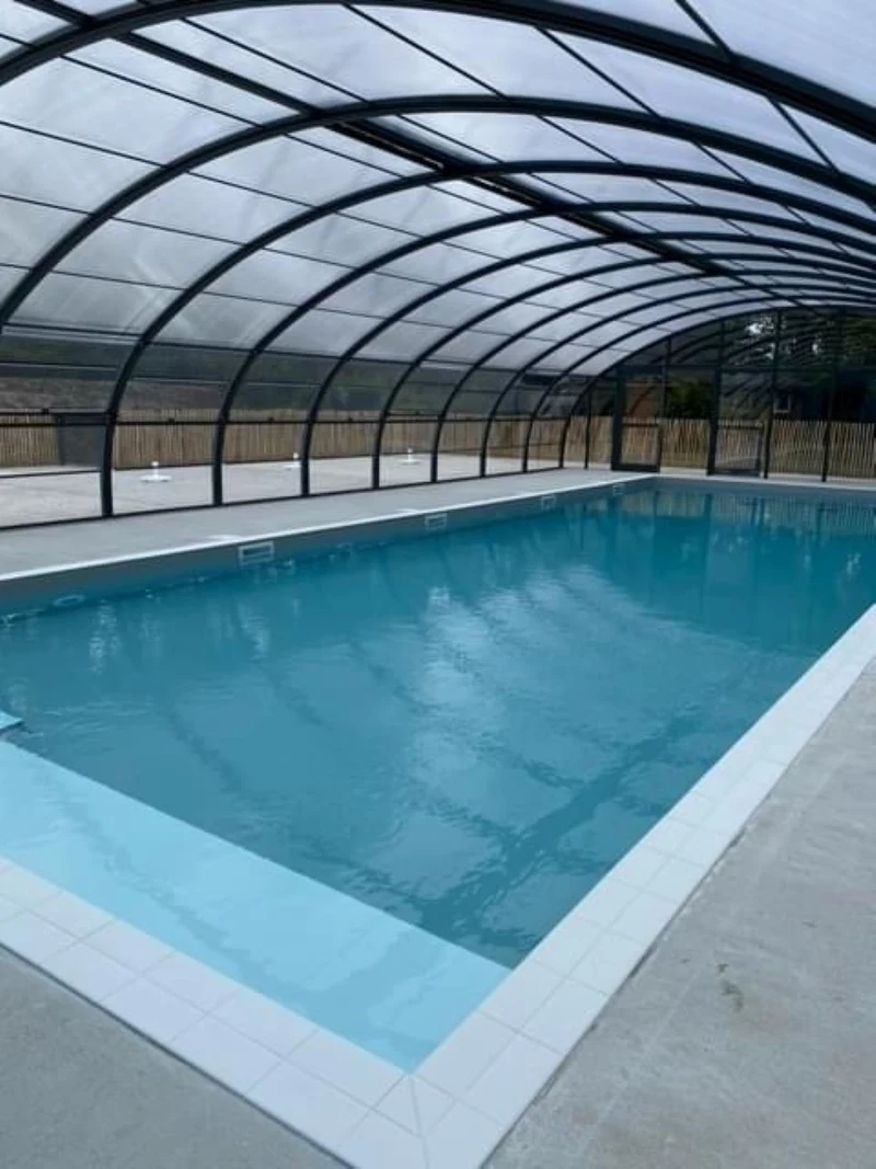 A covered swimming pool with clear water, under a translucent arched roof, surrounded by a concrete deck at Seasonova Du Chêne