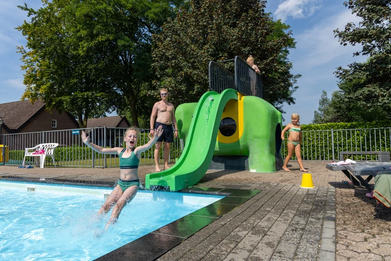 Child slides into a pool, splashing water, with adults and children nearby under sunny skies and trees; at Hoeve de Schaaf