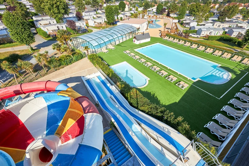 Waterslide curving down into view, overlooking swimming pools and lounge chairs, surrounded by mobile homes and trees at Marvilla Parks La Vallée
