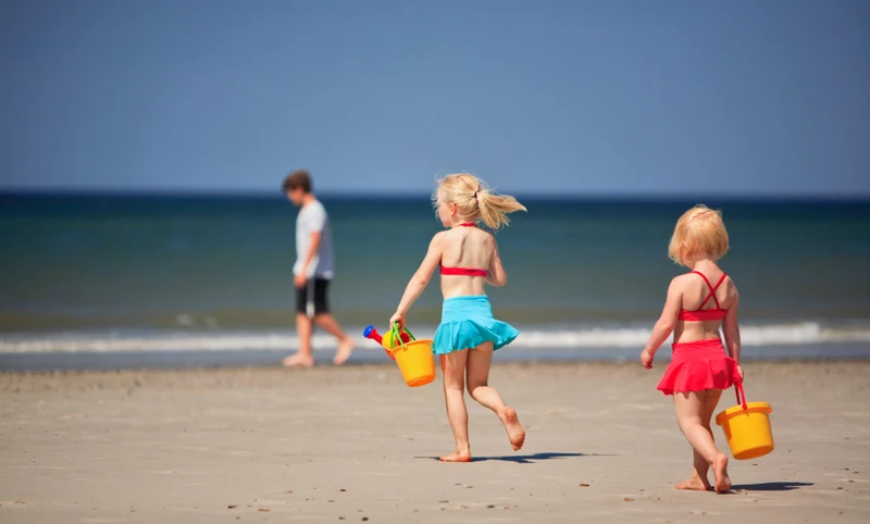 Two children carry yellow buckets, running on a sandy beach with ocean waves in the background at Dancamps Nordsø