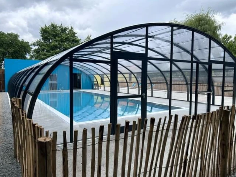 Indoor swimming pool with curved glass roof, surrounded by a wooden fence, under a cloudy sky at Seasonova Du Chêne