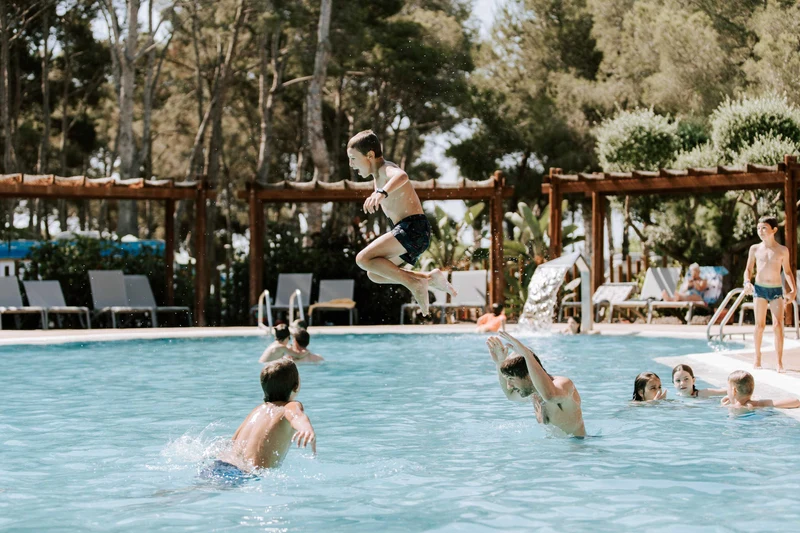 A boy jumps into a pool, surrounded by people swimming and lounging, amidst tall pine trees at Tamarit Beach Resort
