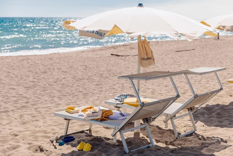 Two lounge chairs under an umbrella, surrounded by beach items, on a sandy shore near the ocean at Club Degli Amici