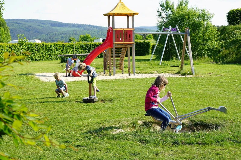 Children playing on playground equipment in a grassy area with a slide, swings, and digging tool at Brilon