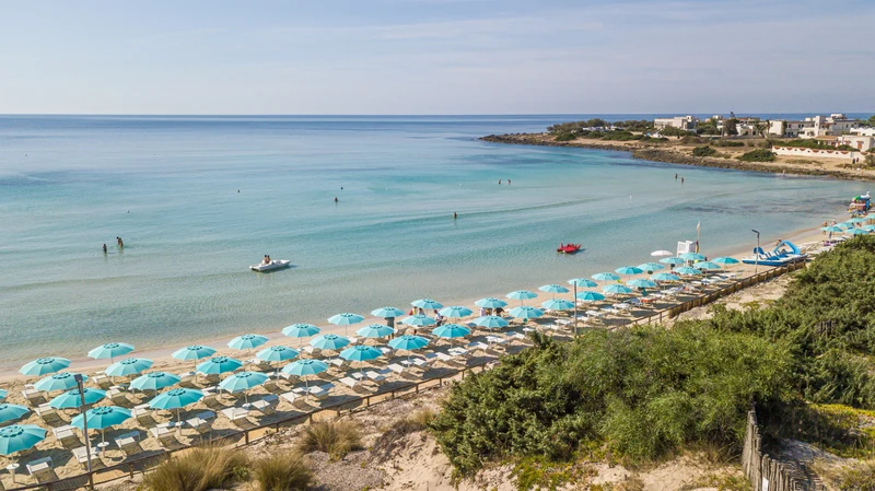 Turquoise umbrellas line sandy beach; people swim and boat in calm, clear sea; coastal buildings in background at Porto Cesareo Camping