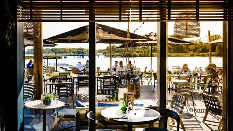 People dining at outdoor restaurant tables under umbrellas near a calm lake, with trees in the distance at Maasresidence Thorn
