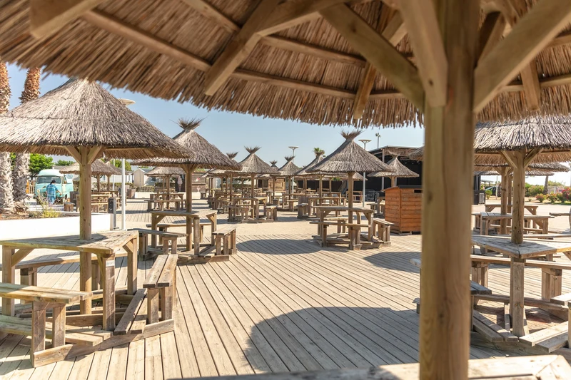Wooden picnic tables with thatched umbrellas on a sunlit deck, surrounded by palm trees at Marvilla Parks Le Castellas