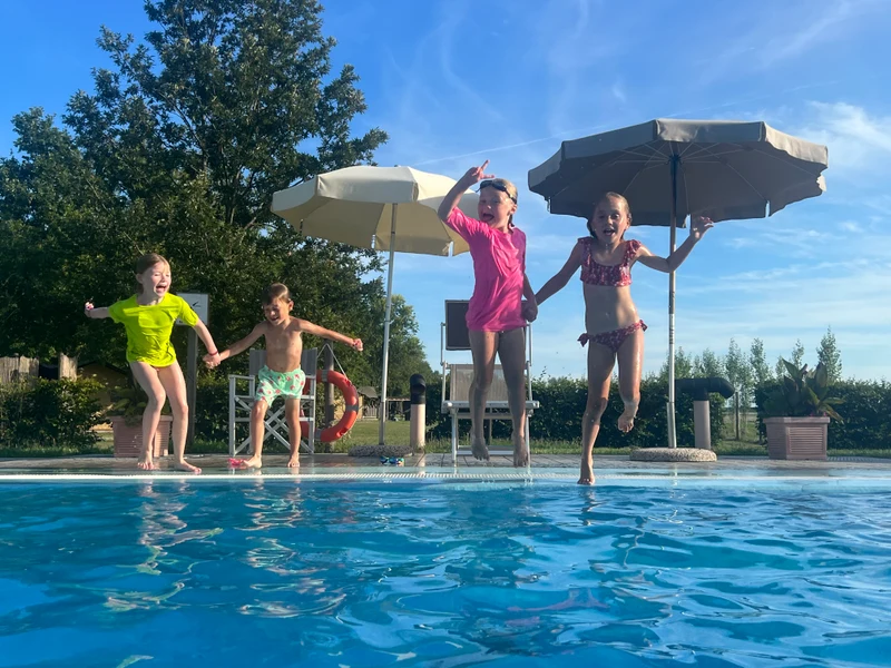 Four children are jumping into a swimming pool, holding hands, with umbrellas and greenery in the background at Agriturismo Tenuta Regina