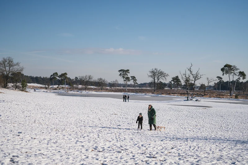 People walking across snow-covered field near frozen pond under clear sky; bare trees and distant forest in background at Marvilla Parks Kaatsheuvel